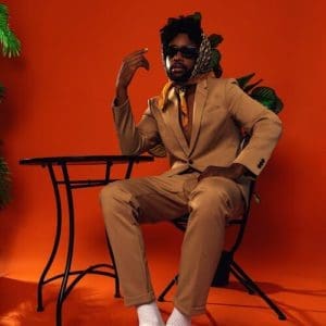 Henry posing sitting in a chair in a tan suit, orange background