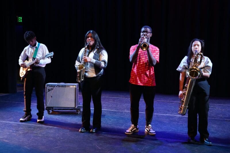 JLG00945 Four musicians stand in a row playing instruments